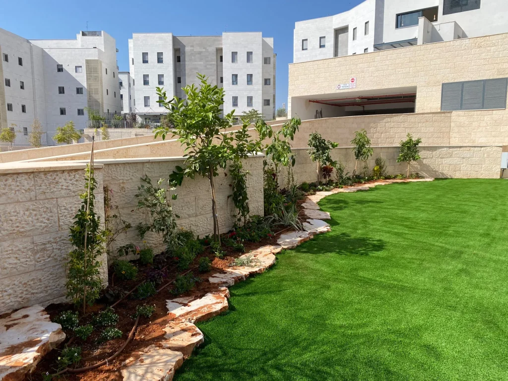 ערוגה ארוכה לאורך קיר אבן עם עצים צעירים, שפת סלעים ודשא סינטטי מול הבית
