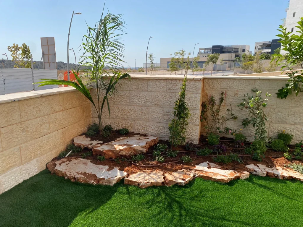 פינת הגינה עם דקל צעיר, שיחים ושפת סלעים בהירים לצד הדשא הסינטטי