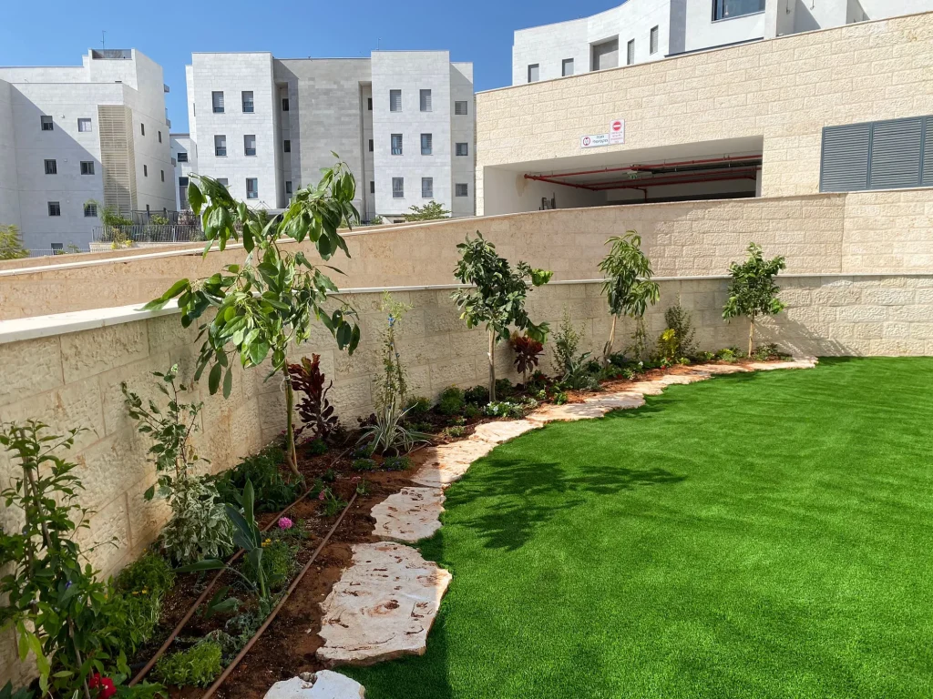 עצים ושיחים בערוגה לאורך הקיר עם קו סלעים מתעקל ודשא סינטטי