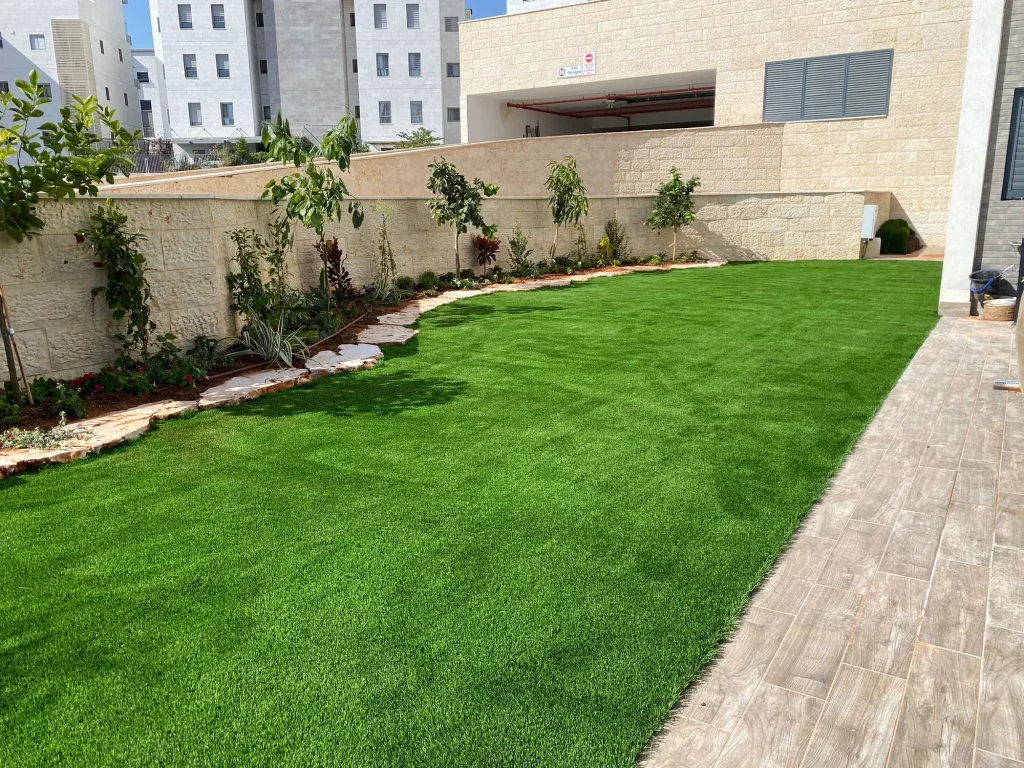 דשא סינטטי לאורך הבית עם ערוגה מתעקלת ושביל ריצוף בצד
