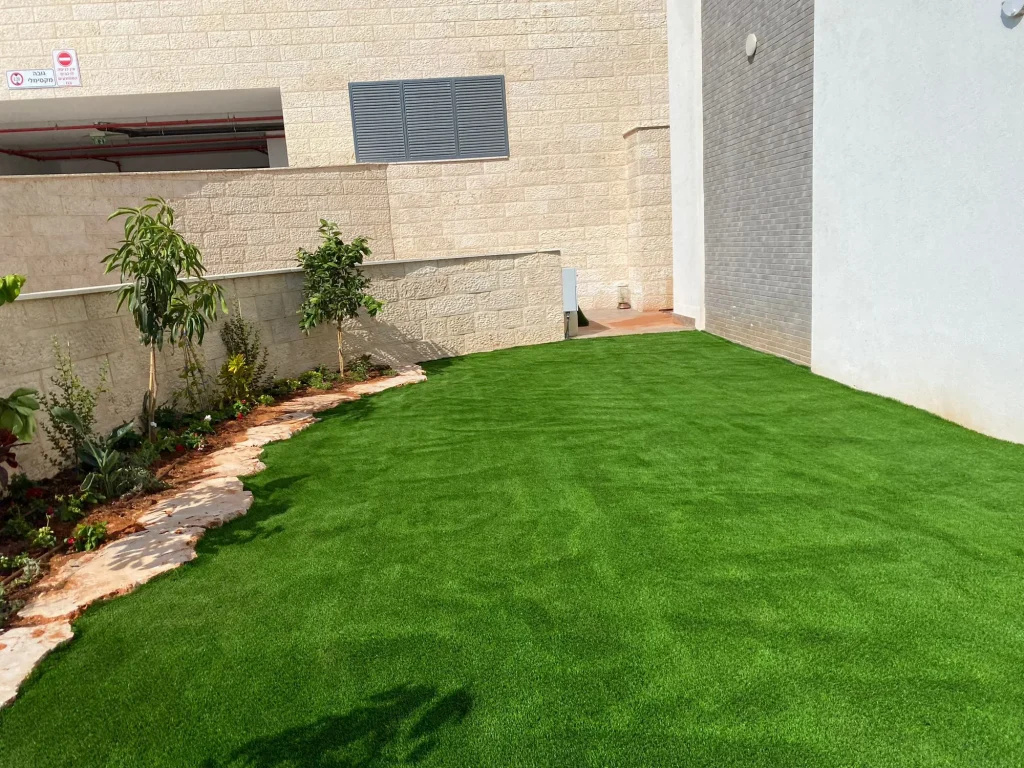 פינת דשא סינטטי ליד הבית עם ערוגת עצים ושפת סלעים