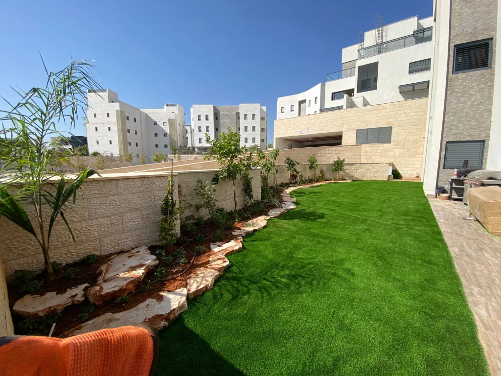 דשא סינטטי לאורך הבית עם ערוגה מתעקלת, סלעים ודקל צעיר