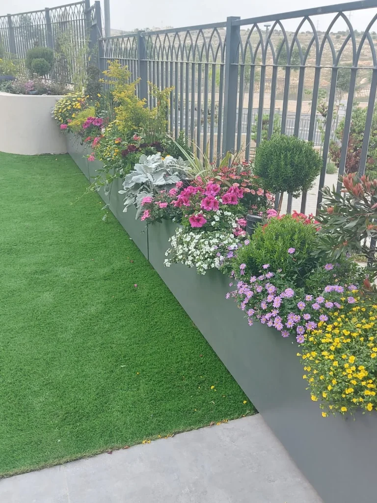 אדניות מתכת אפורות לאורך הגדר שתולות בפרחים צבעוניים לצד דשא סינטטי